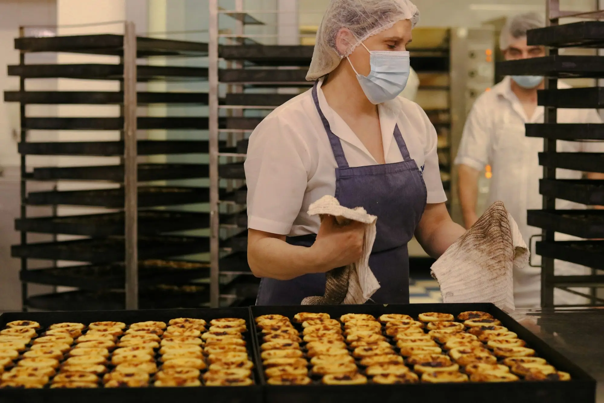 Baker in a hairnet and apron lifting trays of freshly baked pastries beside a trolley of racks in a commercial bakery kitchen